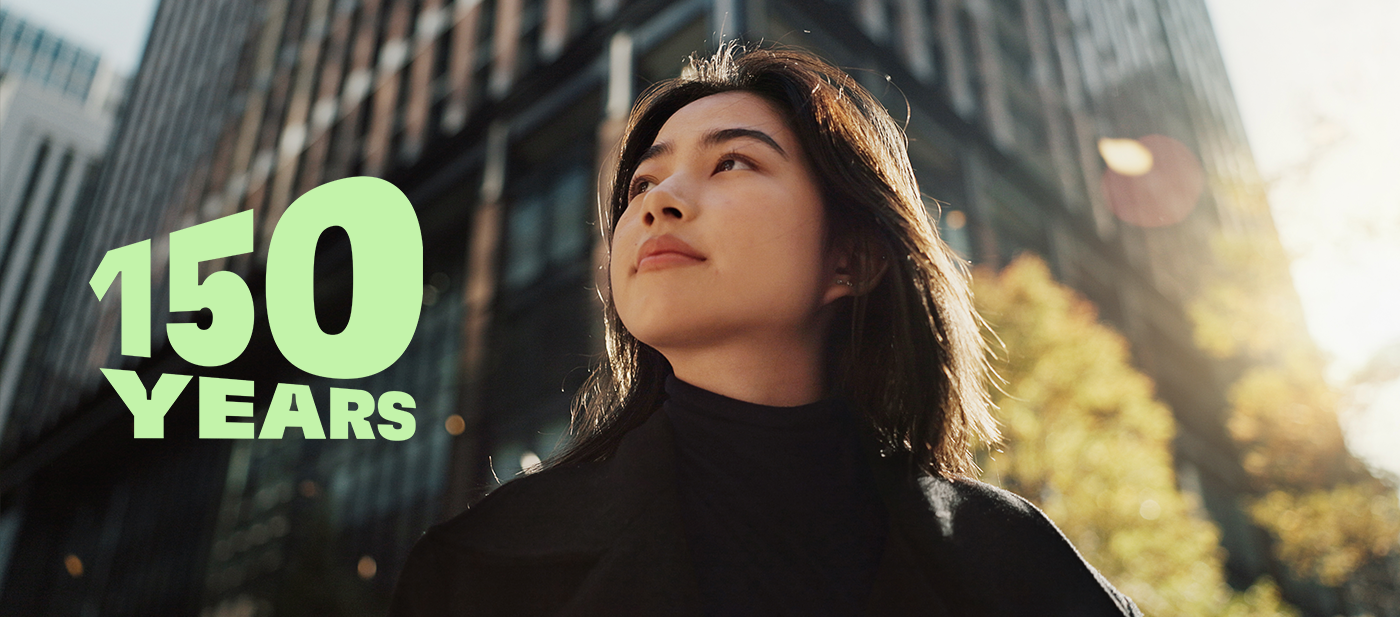 El texto verde dice "150 AÑOS" sobre un paisaje urbano moderno con altos edificios de cristal y árboles otoñales, que simboliza la celebración del 150 aniversario.