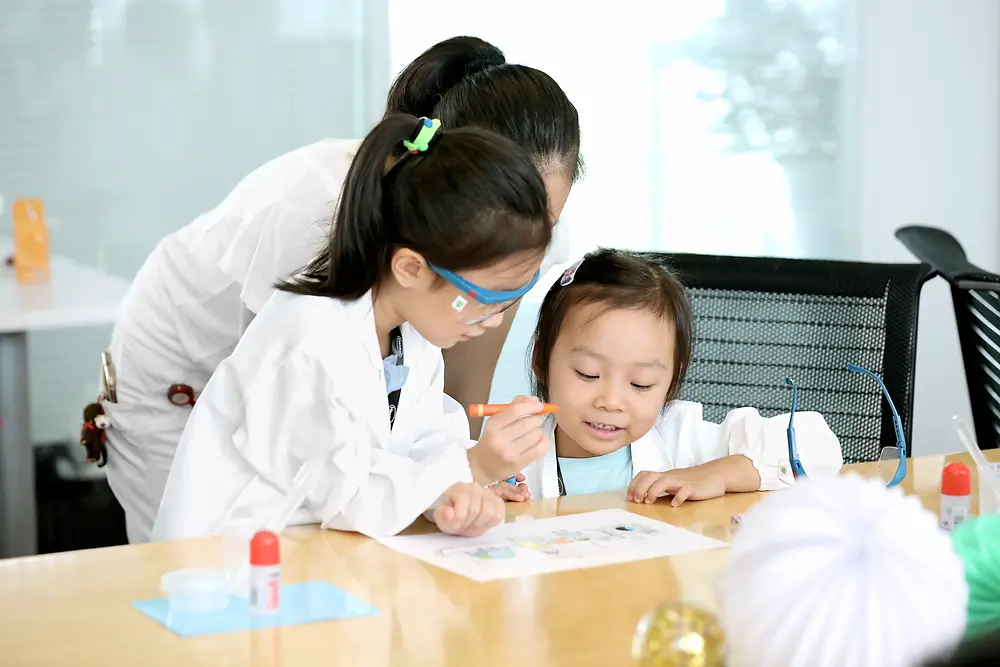 Dos niñas y una mujer con bata de investigación colorean un cuadro en una mesa.