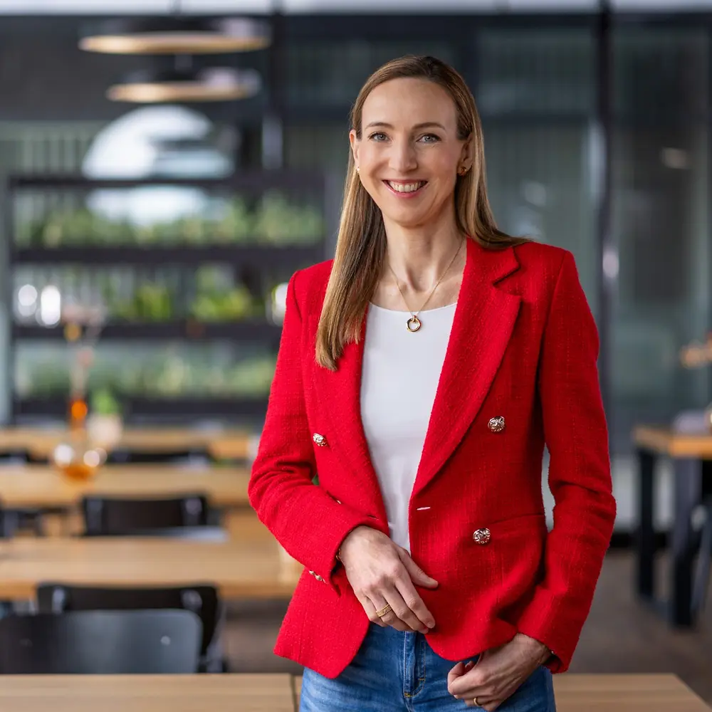 La Dra. Simone Bagel-Trah, vestida con una chaqueta roja brillante y una blusa blanca, de pie en un moderno café con mesas de madera y jarrones decorativos al fondo.