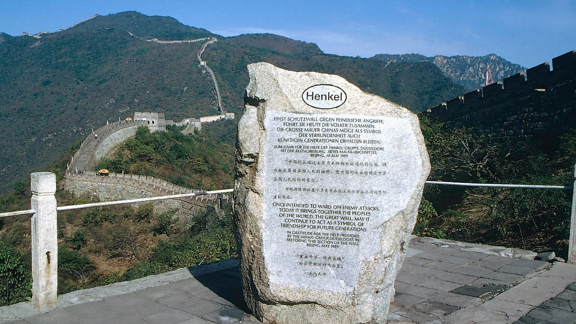 Una lápida conmemorativa en honor al apoyo de Henkel a la restauración de una sección de la Gran Muralla China. Al fondo, la Gran Muralla China serpentea a través de una zona montañosa boscosa.
