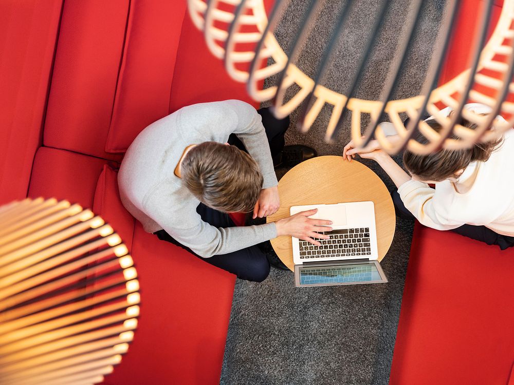 Foto cogida desde arriba con 2 empleados que están sentados en los sofás rojos mirando el portátil en una mesa pequeña. 
